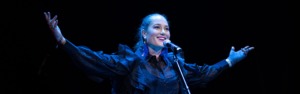 Individual stands in front of a keyboard, on stage, her arms are spread wide and she is smiling. There is a blue stage downlight on her.