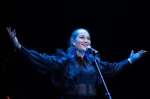 Individual stands in front of a keyboard, on stage, her arms are spread wide and she is smiling. There is a blue stage downlight on her.