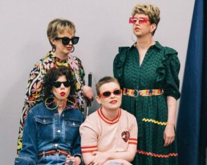 4 individuals with a grey background behind them. Two are standing, two are sitting. They are wearing outfits that include colourful spotted jumpers, denim jackets, dark green dresses and a pale pink jumper. They are all wearing sunglasses.