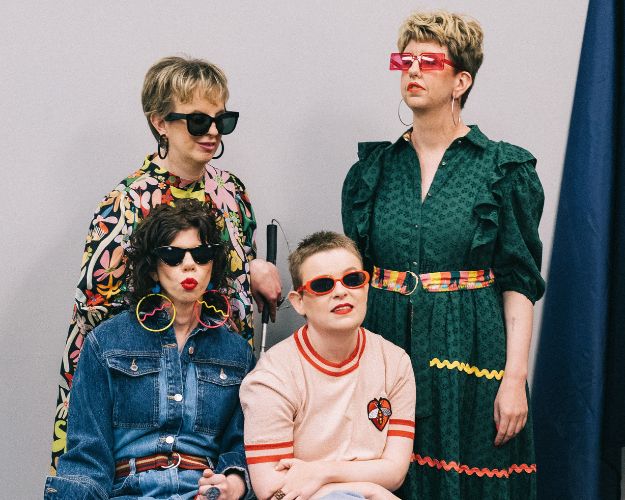 4 individuals with a grey background behind them. Two are standing, two are sitting. They are wearing outfits that include colourful spotted jumpers, denim jackets, dark green dresses and a pale pink jumper. They are all wearing sunglasses.