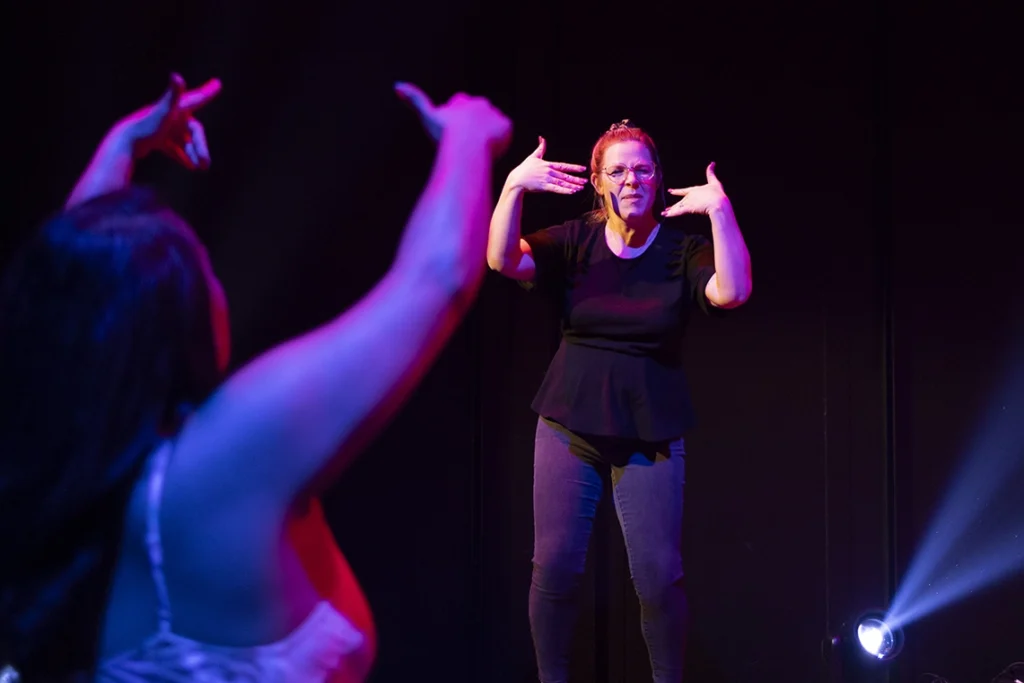 A woman stands in a dark theatre with blue stage lights whilst doing Auslan Interpretation of a performance. A person in the audience is dancing with her.