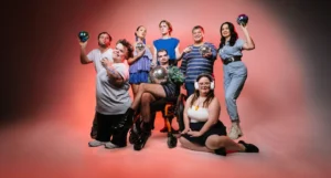 A group of people are standing in front of a white backdrop with dramatic red lights. They are all posing and a few people are holding mirror balls.