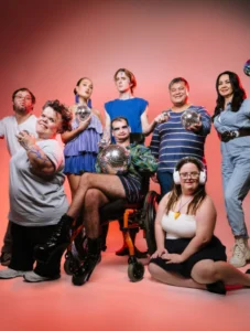 A group of people are standing in front of a white backdrop with dramatic red lights. They are all posing and a few people are holding mirror balls.