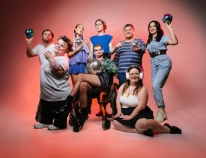 A group of people are standing in front of a white backdrop with dramatic red lights. They are all posing and a few people are holding mirror balls.
