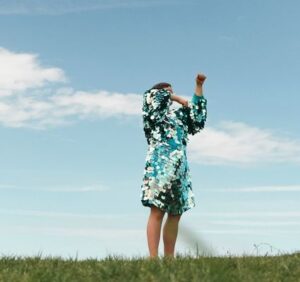 Individual standing on a large open grassy field, blue sky and clouds in the background. They ar wearing a blue green sequin dress with their arms covering their face, they are captured in the middle of movement.