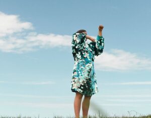 Individual standing on a large open grassy field, blue sky and clouds in the background. They ar wearing a blue green sequin dress with their arms covering their face, they are captured in the middle of movement.
