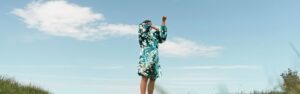 Individual standing on a large open grassy field, blue sky and clouds in the background. They ar wearing a blue green sequin dress with their arms covering their face, they are captured in the middle of movement.