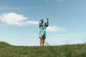 Individual standing on a large open grassy field, blue sky and clouds in the background. They ar wearing a blue green sequin dress with their arms covering their face, they are captured in the middle of movement.