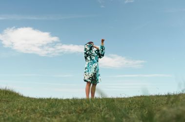 Individual standing on a large open grassy field, blue sky and clouds in the background. They ar wearing a blue green sequin dress with their arms covering their face, they are captured in the middle of movement.